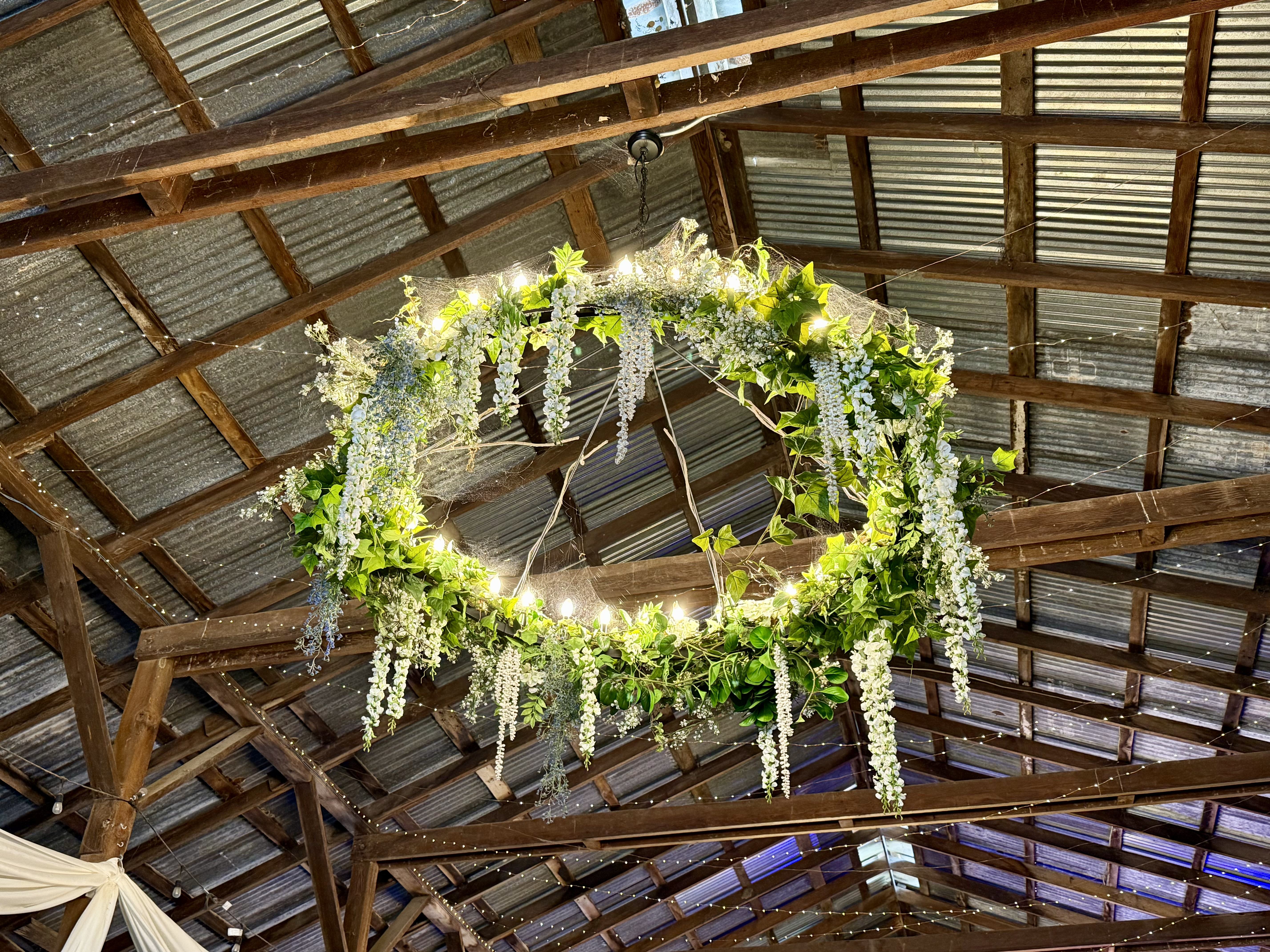 A chandelier in the barn, covered in green leaves and some white flowers, looking like it’s intended for weddings (because it is)